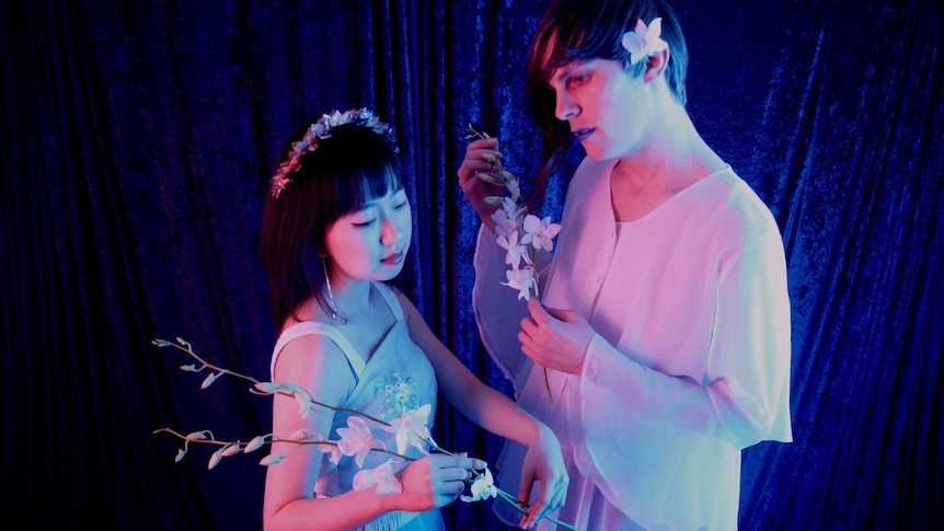 Two people dressed in white stand holding white flowers. Behind them is a blue curtain and  blue light reflects on them.