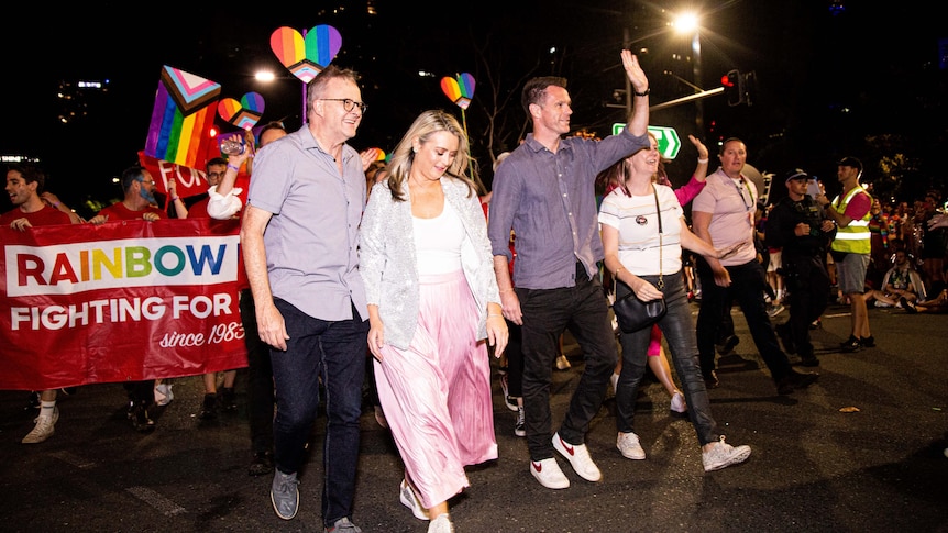 Anthony Albanese walks with others in front of a banner and people holding rainbow flags.