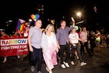 Anthony Albanese walks with others in front of a banner and people holding rainbow flags.