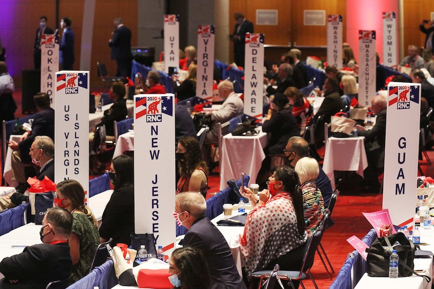 A group of people wearing masks sit at tables with signs printed with a state on it in a room.