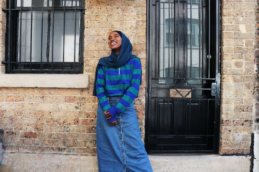 Hijabi model Miski Omar poses in a long denim skirt in front of brick house to depict modest fashion trend.
