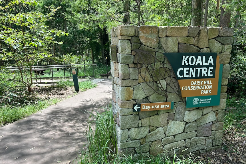 A picture of a sign that says 'Koala Centre: Daisy Hill Conservation Park', with grass, footpath, and trees in the background.