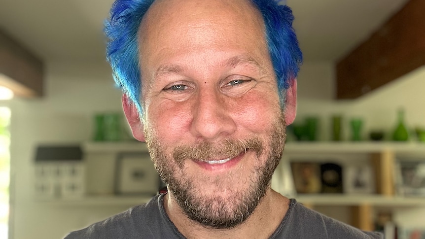 A man with blue hair and brown beard smiles in a close up of his face.