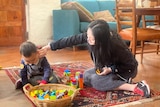 A young Asian woman sits on a colourful rug and reaches out to a baby playing with toys.