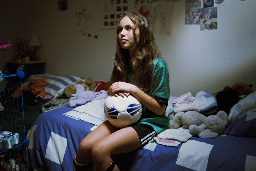 Young female soccer player sits in her bedroom holding a ball