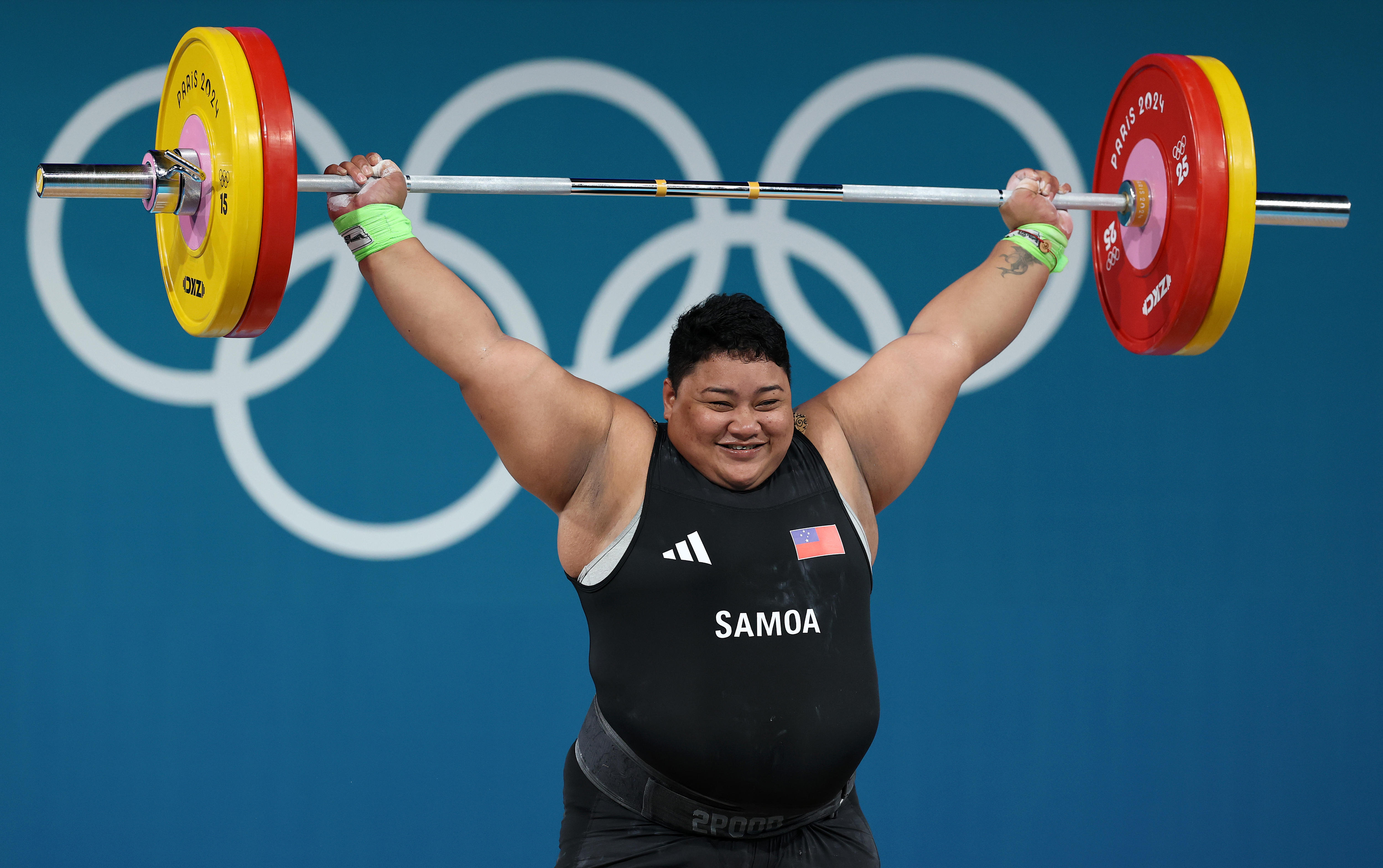 Uma mulher samoana segura uma barra com pesos em cada extremidade acima da cabeça e sorri.