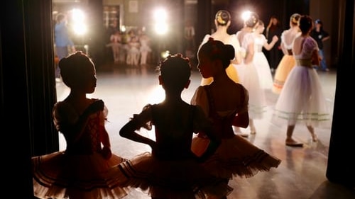 Young ballet dancers are silhouetted as they wait in the wings to go on stage where other dancers can be seen
