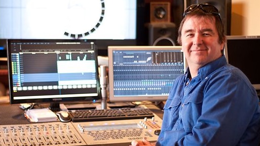man in blue shirt smiling with hands clasped with audio desk and computer screens in the background