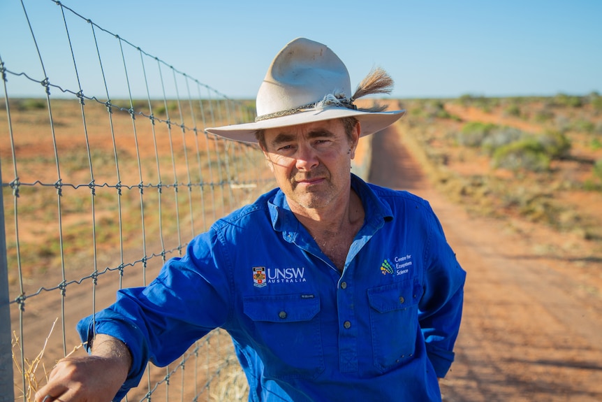 A man in a blue shirt and wide-brimmed hat standing by a fence in the outback