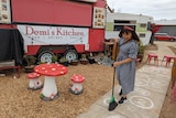 A woman with short black hair with a bow in it and gingham dress is sweeping outside her red food van. 