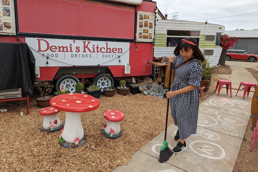 A woman with short black hair with a bow in it and gingham dress is sweeping outside her red food van. 