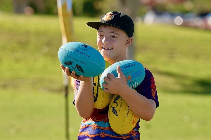 Boy wearing cap smiles and holds four footballs on a oval in Broome
