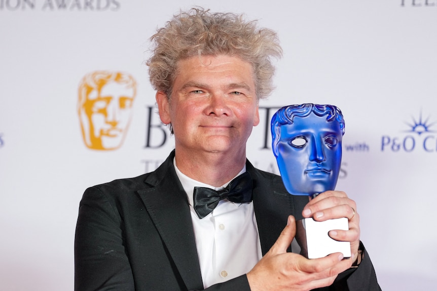 A middle-aged man in a suit and bow tie holds aloft a BAFTAS award and smiles gently.