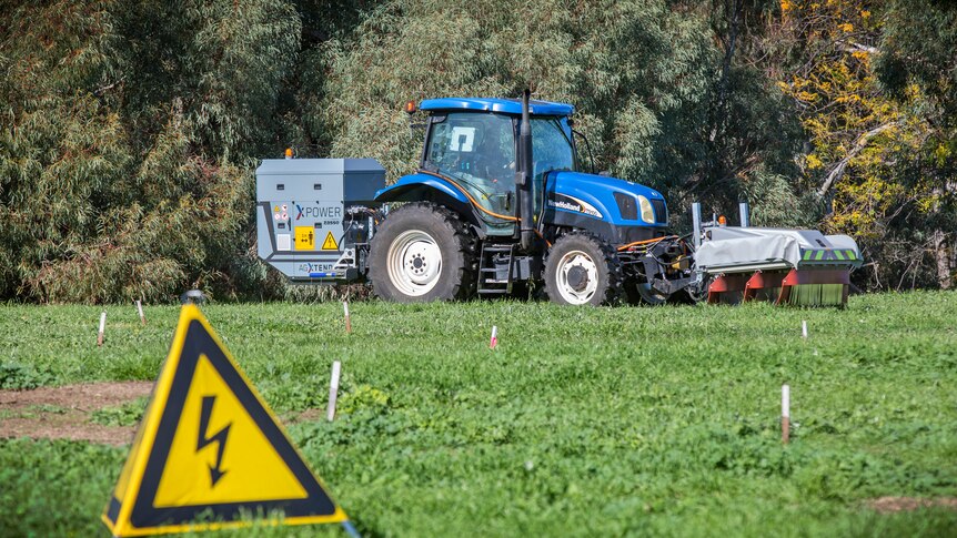 A warning sign sits on grass in front of a tractor.