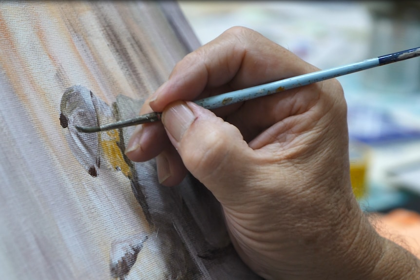 A hand holding a paint brush, and putting paint strokes on a canvas.