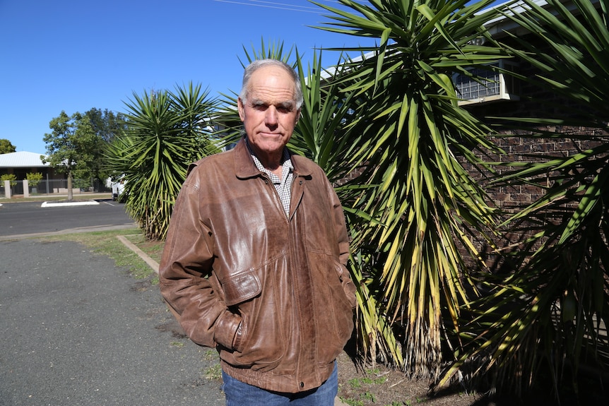 A man in a in a brown leather jacket stands outside with his hands in his pockets