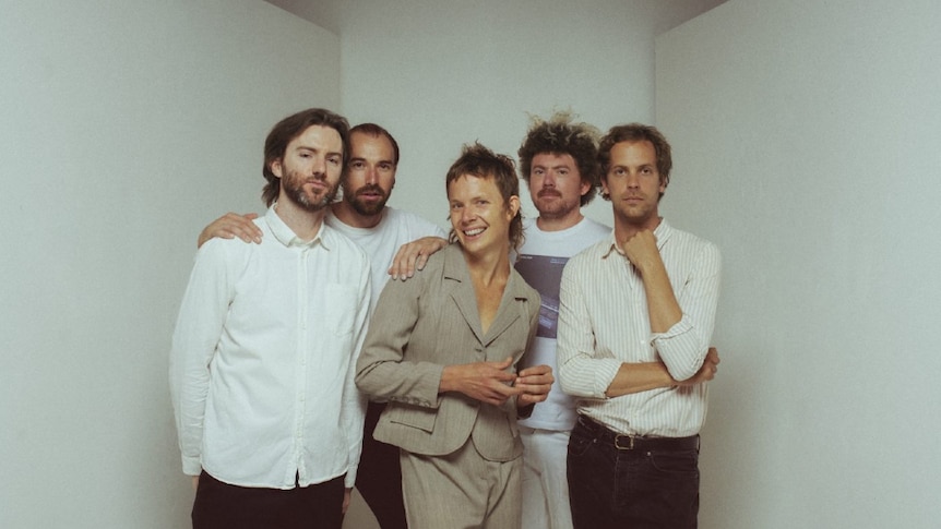 Five men, three wearing white shirts, one in beige suit, and one in striped shirt and dark pants