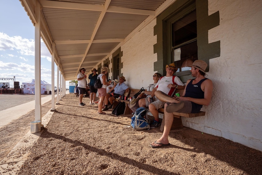 Men in shorts, tee,hat sitting, some standing, in a deep verandah,  cream wall with brown detailing on the windows.