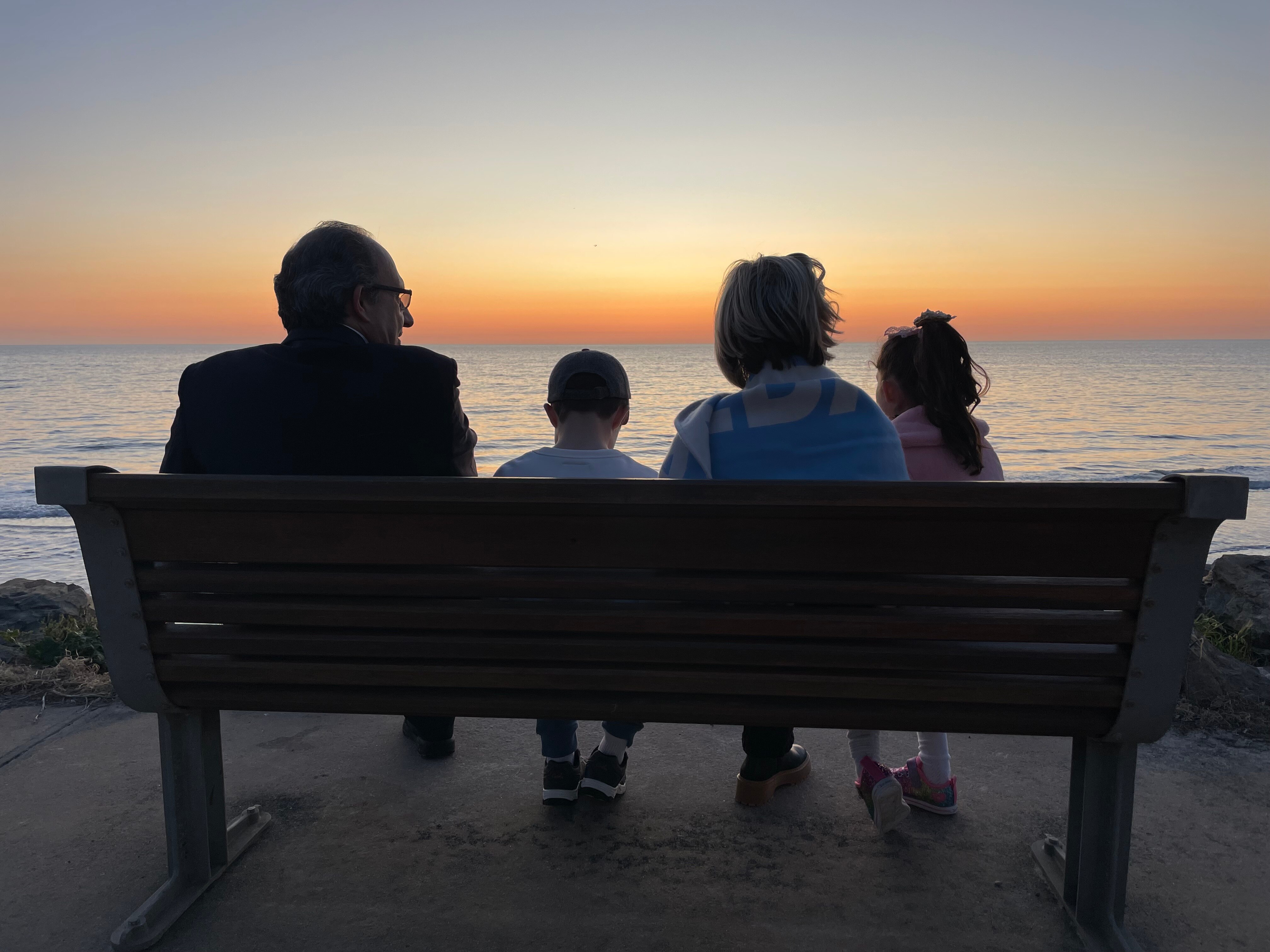 A family of two children and woman silhouette as they sit on a bench looking at beach sunset