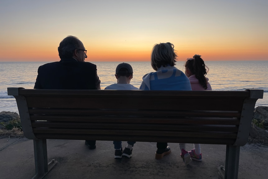A family of two children and woman silhouette as they sit on a bench looking at beach sunset