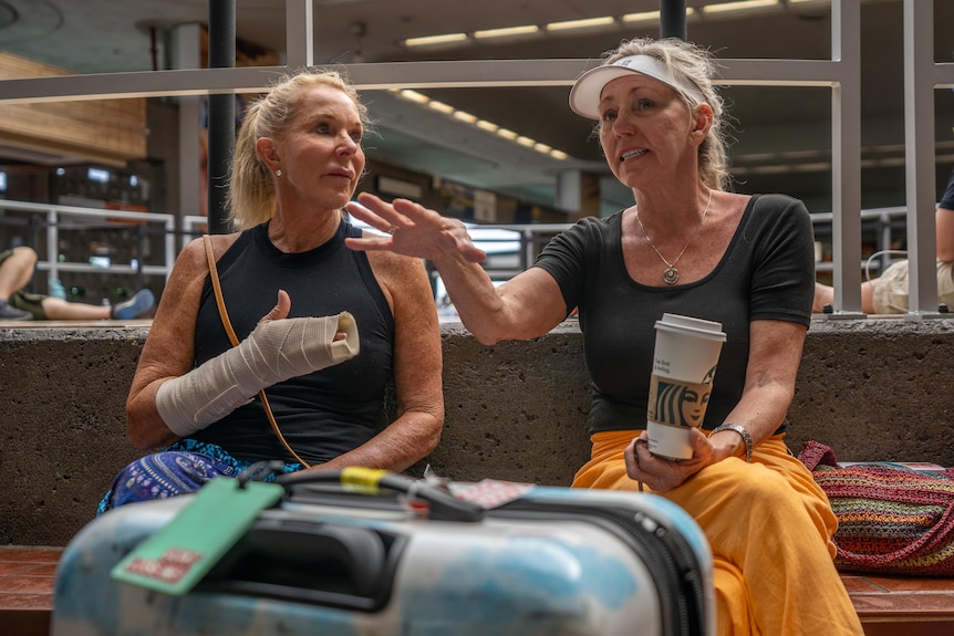 Two women sit on an airport bench. One has her hand in plaster. The other holds a coffee. There are bags beside them.