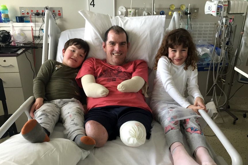 A man with all limbs amputated lies in a hospital bed, smiling with a young boy and girl next to him in the bed