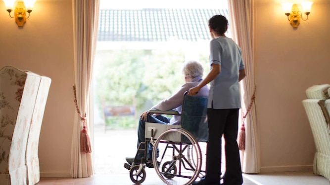 An aged care worker with a resident in wheelchair sitting at window.