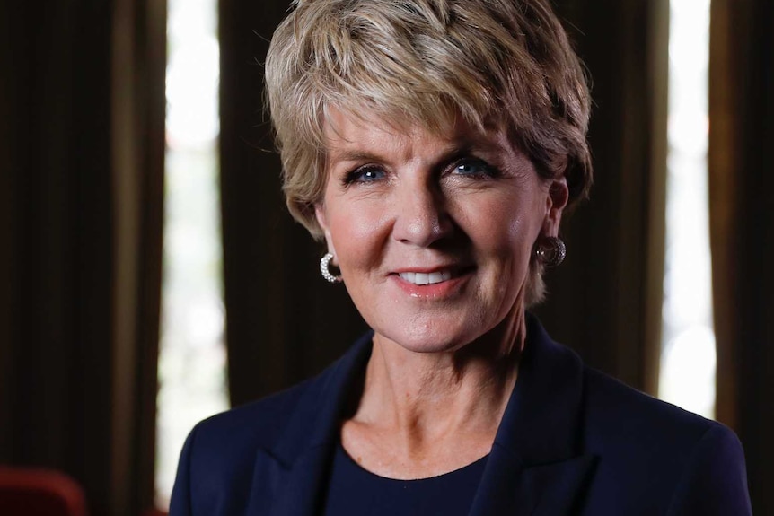 Ms Bishop is smiling and facing the camera, in a navy blazer. The background is dark, with light peering in through windows.