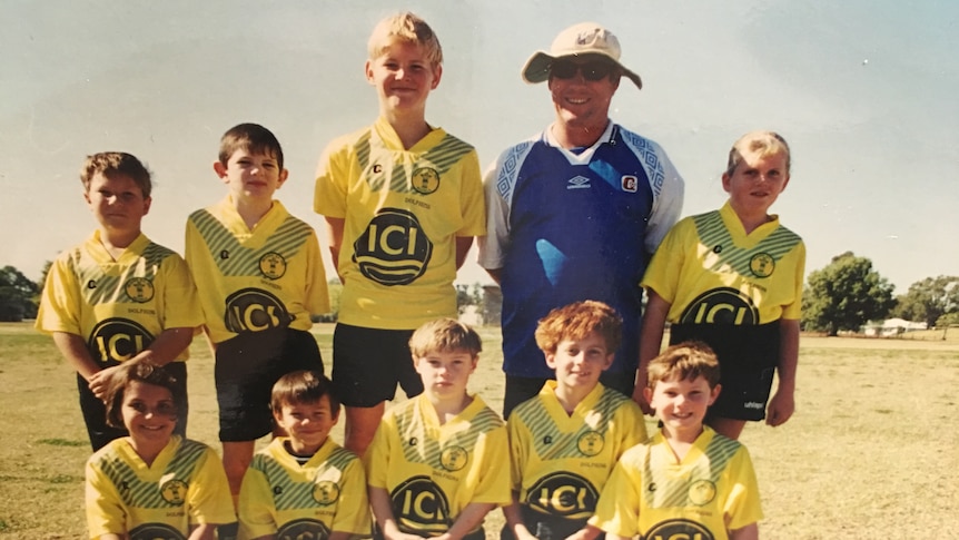 Nine boys in yellow jerseys next to their coach in an outdoor field