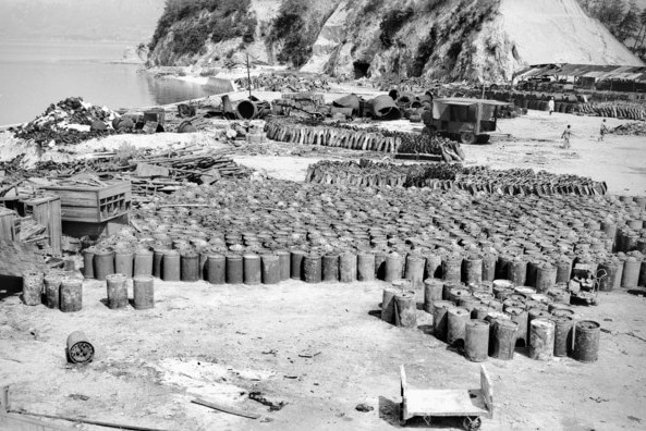 Black and white photo of dozens of barrels against dilapidated wooden structures next to a beach.
