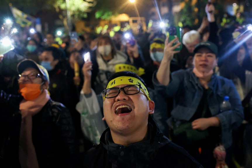 Protesters wearing yellow headbands gather and sing songs, with many holding mobile phones aloft.