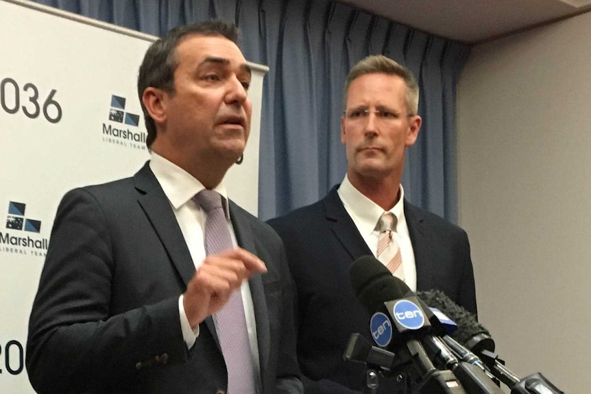 Two men in suits stand in front of a podium.  One is speaking while the other watches.