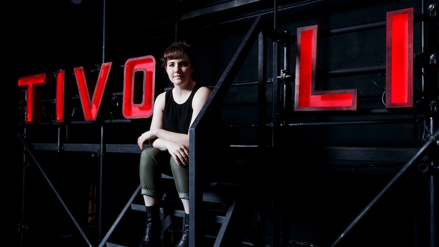 Young woman in black tank top and army-green pants sitting on set with large light-up letters spelling TIVOLI behind her in red.