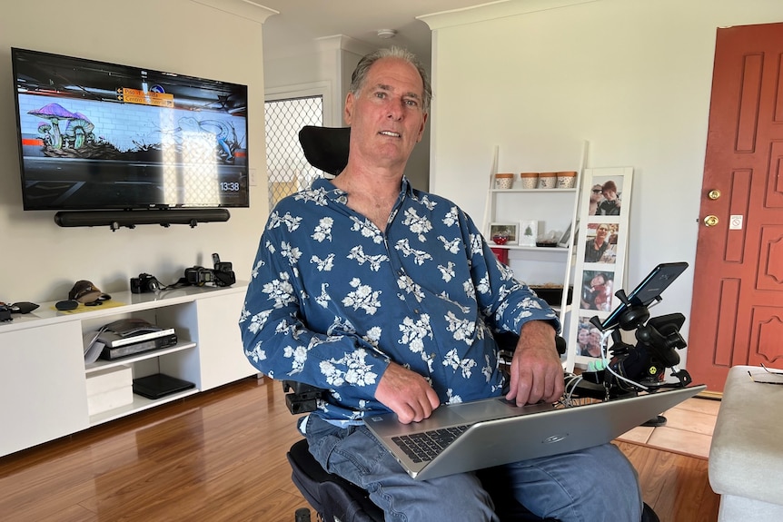 A man sits in an electric wheelchair in his loungeroom with a computer on his lap. 