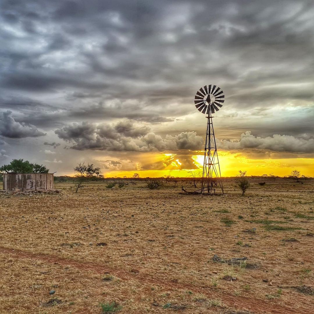 A photograph of a windmill on an outback station, golden sun rays are shining through the grey clouds on the horizon.