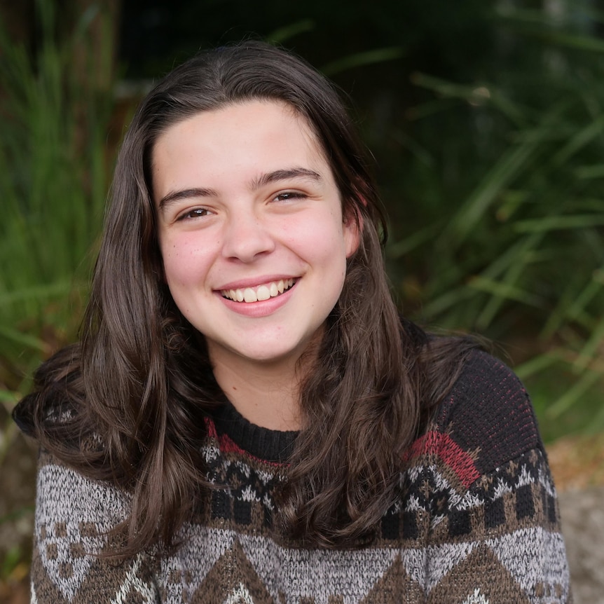 young girl with brown medium length hair smiles at the camera wearing a patterned winter jumper. 