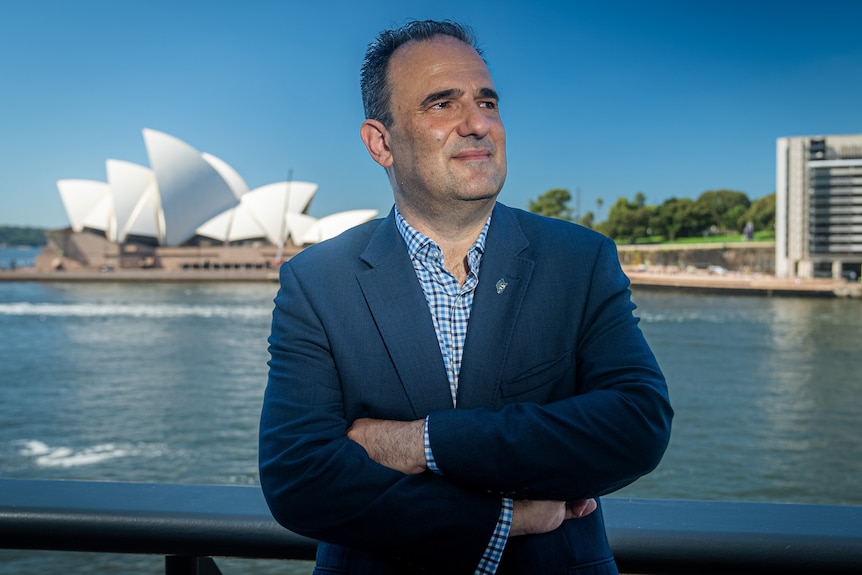 A man in a blue jacket stands in front of the Sydney Opera House