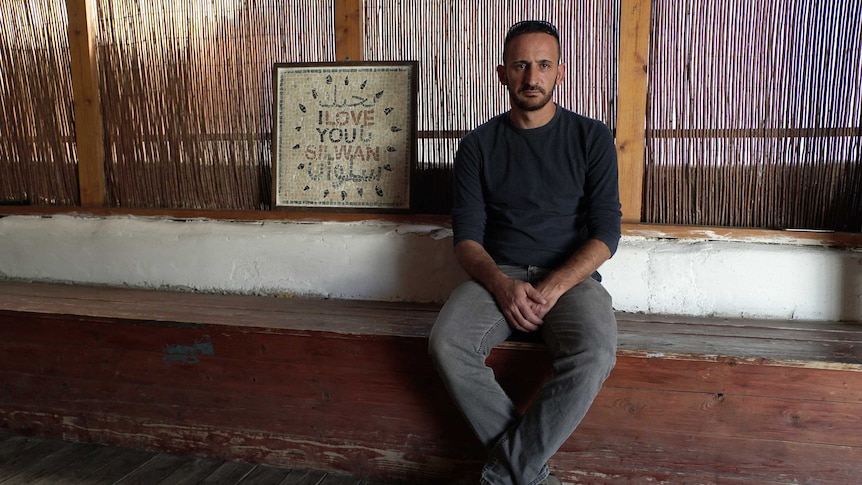 A bearded man sits next to a sign that reads "I love you Silwan" with Arabic text above and below