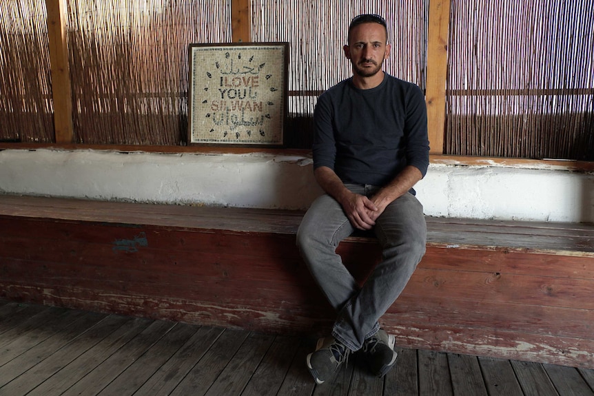 A bearded man sits next to a sign that reads "I love you Silwan" with Arabic text above and below