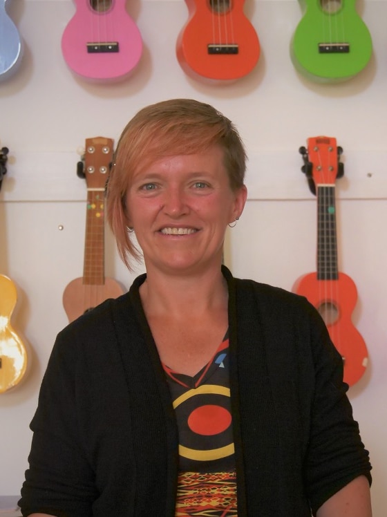 A woman stands in front of several ukuleles which are hanging on the wall