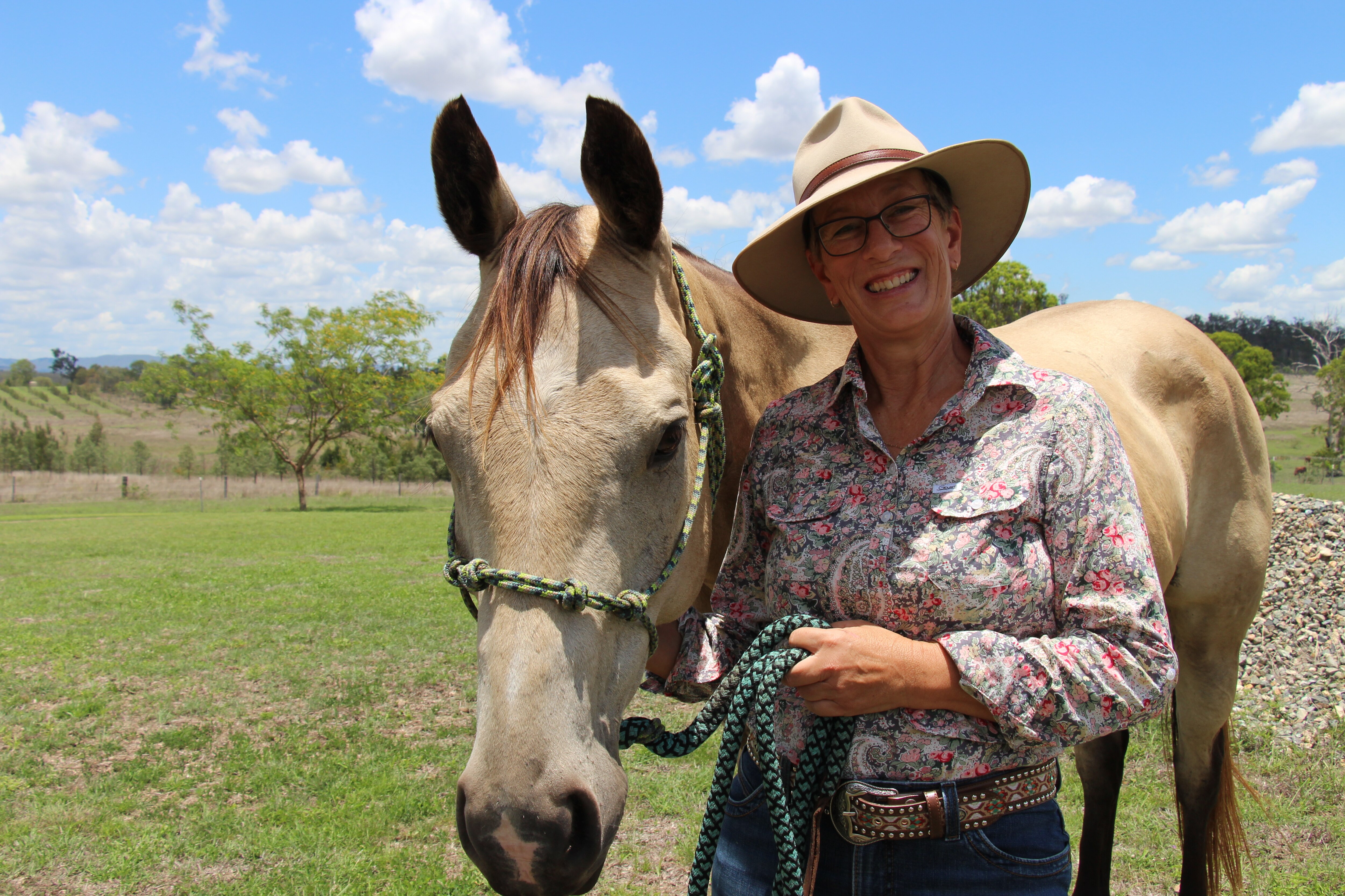 A woman in a button-up shirt and hat holds a halter rope, a beige-coloured horse beside her