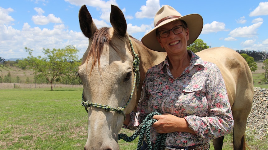 A woman in a button-up shirt and hat holds a halter rope, a beige-coloured horse beside her