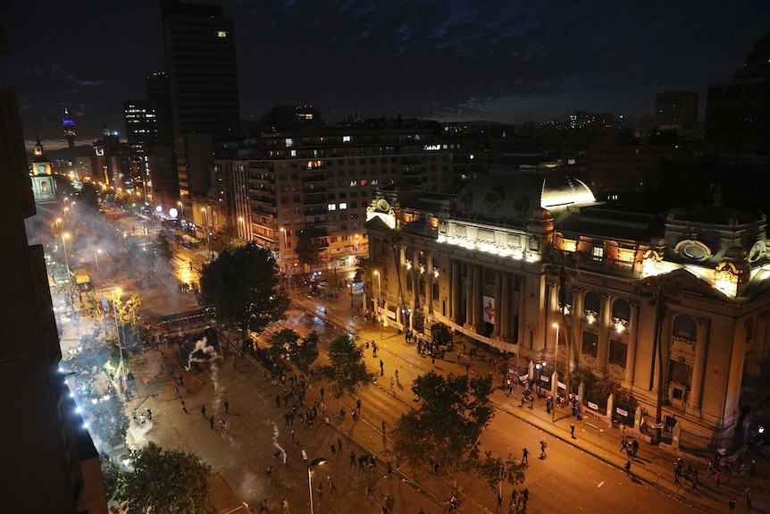 A shot of a wide boulevard in Santiago at night, the street aglow and plumes of smoke drifting up from tear gas canisters