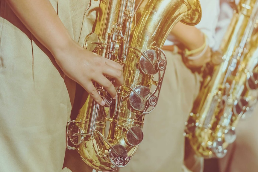 A close-up shot of a saxophone and an arm holding it, with fingers pressing buttons. In background are other blurred saxophones.
