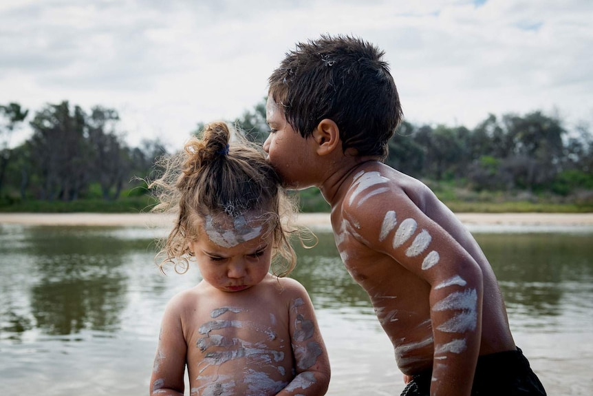 A young boy kisses his sister, they are Indigenous and have clay lines painted on their bodies and faces