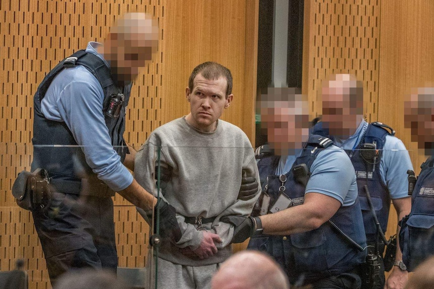 Brenton Tarrant in handcuffs being seated in a courtroom by cops