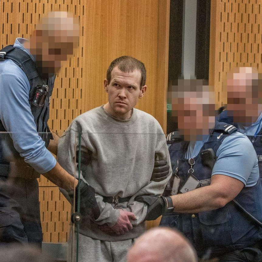 Brenton Tarrant in handcuffs being seated in a courtroom by cops