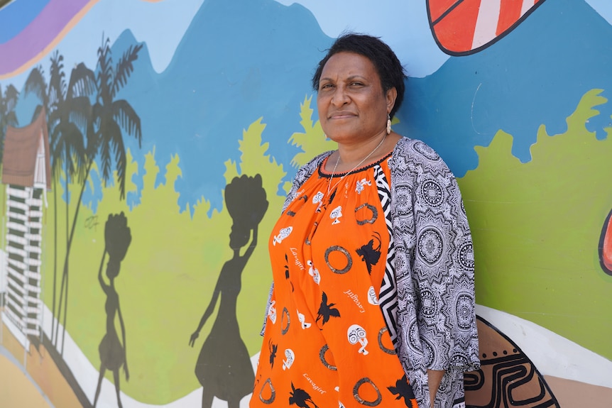 A woman wearing an orange dress with a grey jacket stands against a wall coloured in green and blue.