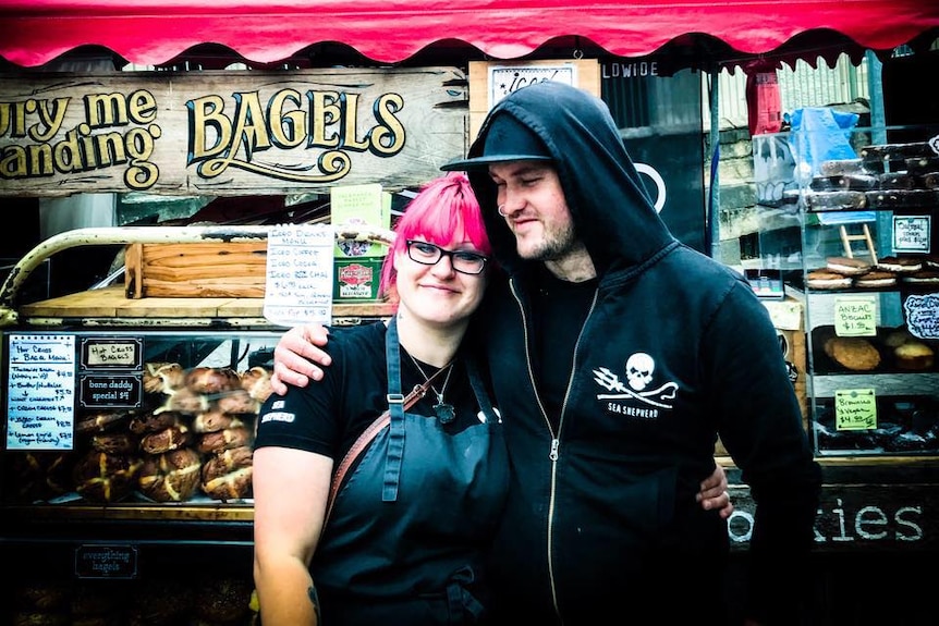 Two people stand in front of a bagel stand.