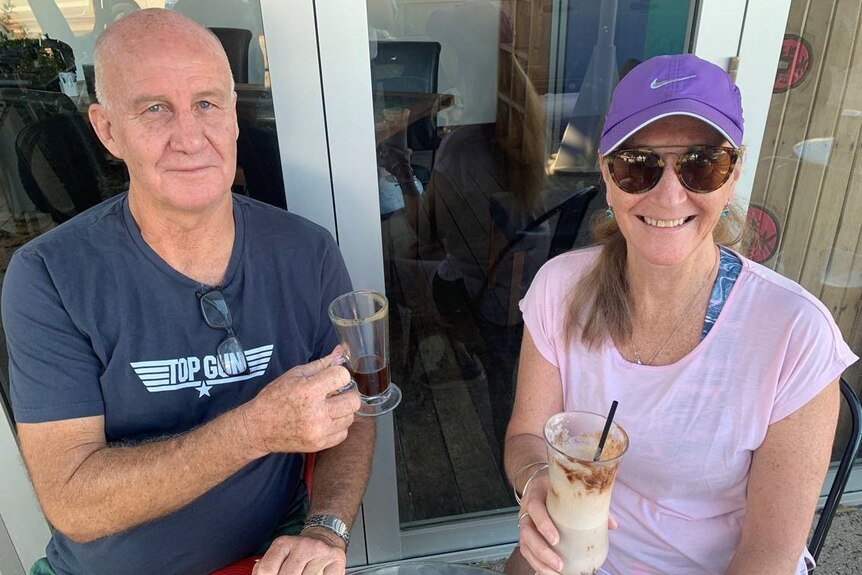 A woman and a man, who are both wearing casual clothes, smile as they sit near a cafe window with drinks.